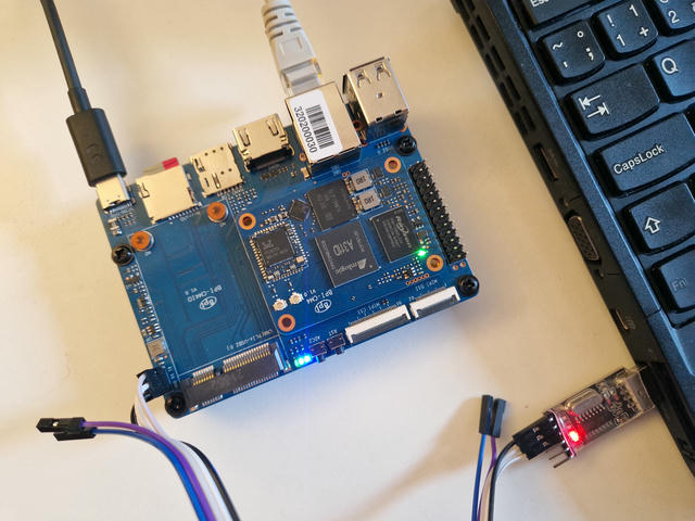 Blue pcb on a white desk, it's the Banana Pi IO board and Compute Module 4.
Along the top edge are a USB C power input, micro SD card, empty SIM slot and HDMI connector, RJ45 ethernet and two empty USB A ports.
Right half of the IO board is covered by the SoM. It has an Amlogic A311D SoC, Rayson RAM, Samsung e-MMC, Realtek Ethernet controller and a Realtek Wifi and Bluetooth stamp module with two U.FL antena connectors next to it.
Left half of the IO board has a vacant mini-PCI Express connector.
Along the right edge is the typical Raspberry Pi 2.54mm spacing pin header.
Along the bottom edge are two flat-flex cable connectors labeled MIPI DSI, and MIPI CSI (for display and camera), two buttons, one Reset and one labeled "ADC2" which I don't know what does yet, and some indicator LEDs.
Along the left edge is 3 pin UART header which is connected to a cheap USB UART adapter connected to a black laptop that's mostly off screen.