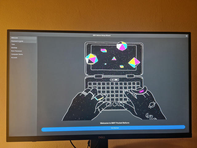 Desktop monitor showing the MNT Pocket setup wizard. The first screen contains a stylized drawing of the Pocket Reform and some hands typing on it. The drawing has black background with white outlines and colorful abstract shapes colored in with cyan, magenta and yellow solid colors.