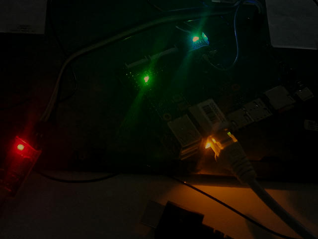 Grainy picture of the Banana Pi board, it's dark, the LEDs on the board itself, on the ethernet and on the UART adapter are green, blue, yellow and red, like winter holiday lights.